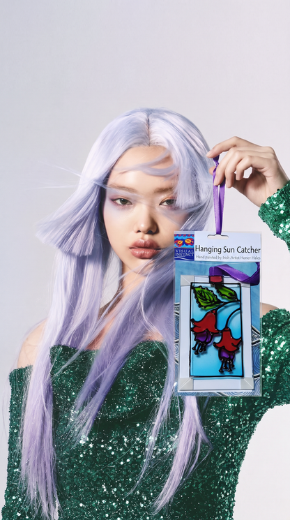 A model with long lavender hair holds up a packaged stained glass hanging sun catcher. The sun catcher features a colorful design of red flowers and green leaves on a blue background.