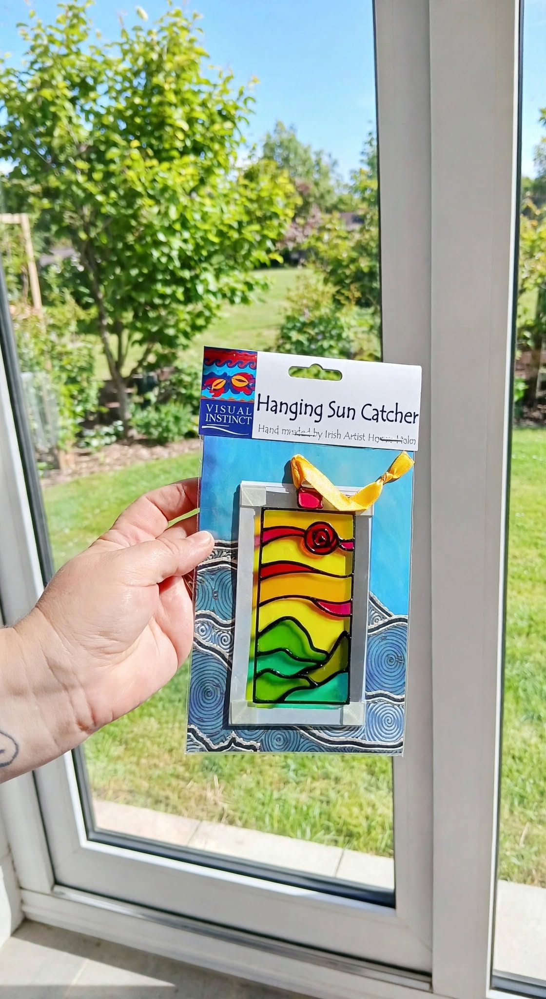A hand holds a packaged hanging sun catcher in front of a window, with a garden visible outside. The sun catcher has a colorful stained-glass-style design with yellow, green, red, and blue sections.