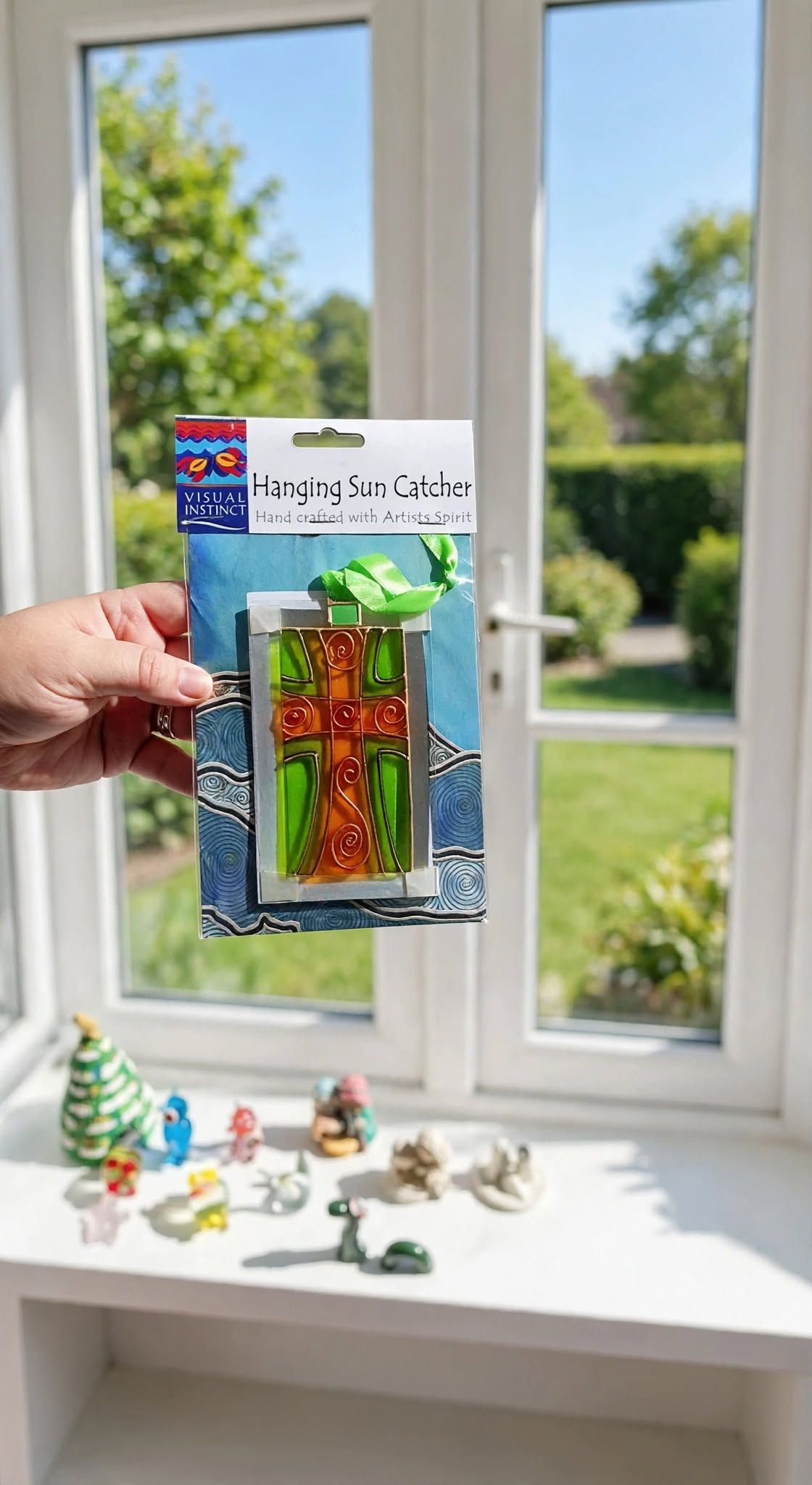 A hand holds a packaged glass sun catcher featuring a green and orange tree design with a blue and white patterned backing, set against a bright window with a garden view.