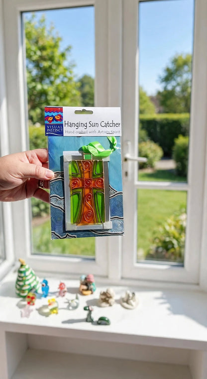 A hand holds a packaged glass sun catcher featuring a green and orange tree design with a blue and white patterned backing, set against a bright window with a garden view.