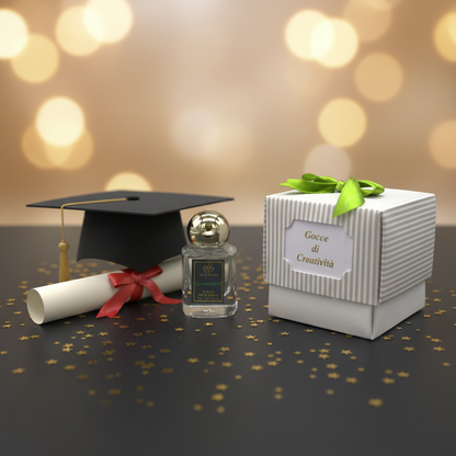 A graduation-themed fragrance gift set featuring a small clear glass perfume bottle with a gold cap, a black graduation cap with a gold tassel, a rolled diploma tied with a red ribbon, and a white striped gift box with a green bow and a label reading 'Gocce di Creatività'. The scene is set on a dark surface with scattered gold confetti and a warm golden bokeh background.