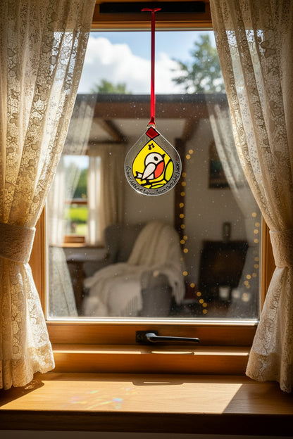A stained glass hanging ornament shaped like a teardrop, featuring a bird with a red breast on a yellow background, bordered by the hand-written quote 'Robyns Appeare When Loves Chris Are Near' in black script, suspended by a red ribbon against a white wall.
