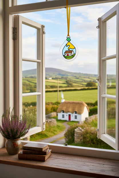 A teardrop-shaped stained glass ornament hanging from a yellow ribbon. The ornament features a hand-painted scene of a small white house with a brown roof, set against a light blue sky, yellow sun, and green grass, all outlined in black glass.
