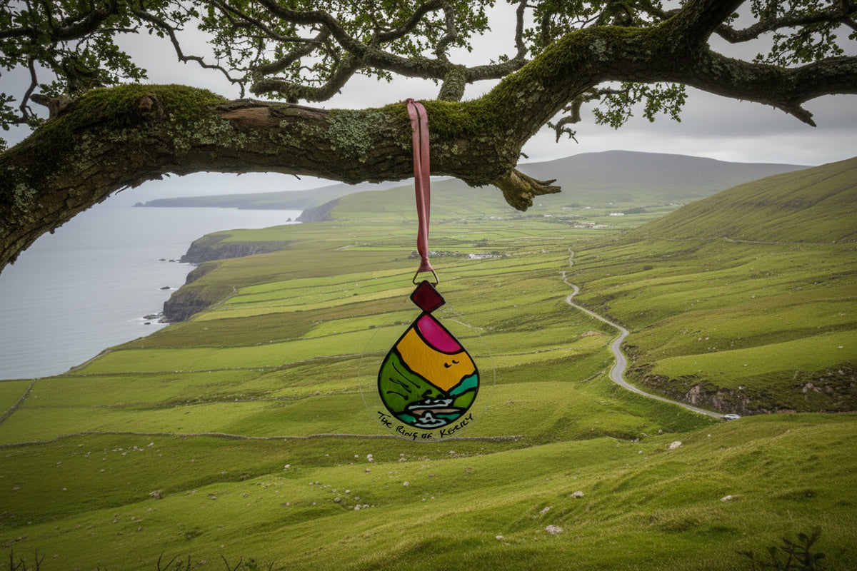 A teardrop-shaped clear glass ornament featuring a colorful stained-glass style illustration of a landscape with green hills, a yellow sun, pink sky, and a boat. The ornament hangs from a pink ribbon and has the text 'The Ring of Kerry' written at the bottom.