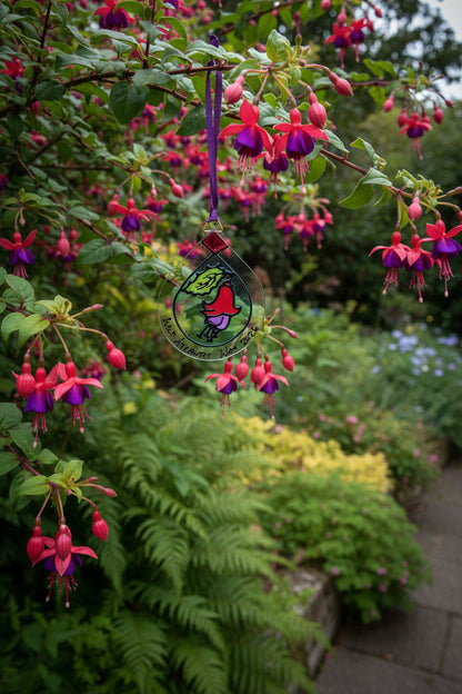 A handcrafted stained glass ornament shaped like a teardrop, featuring a whimsical cartoon character and the text 'Med Attitude War 2026', hanging from a purple ribbon against a textured wall.