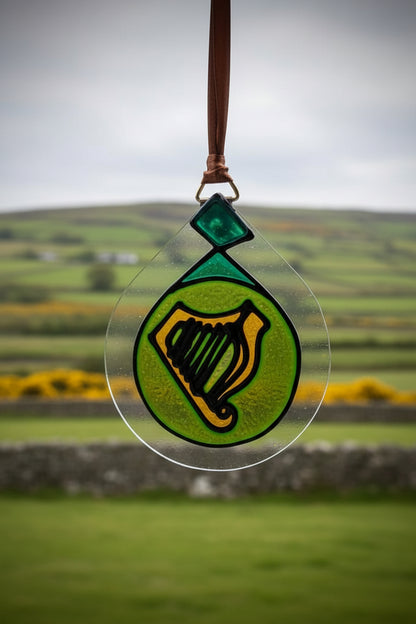 A stained glass ornament featuring a green harp on a bright green circular background, suspended by a brown ribbon against a white wall.