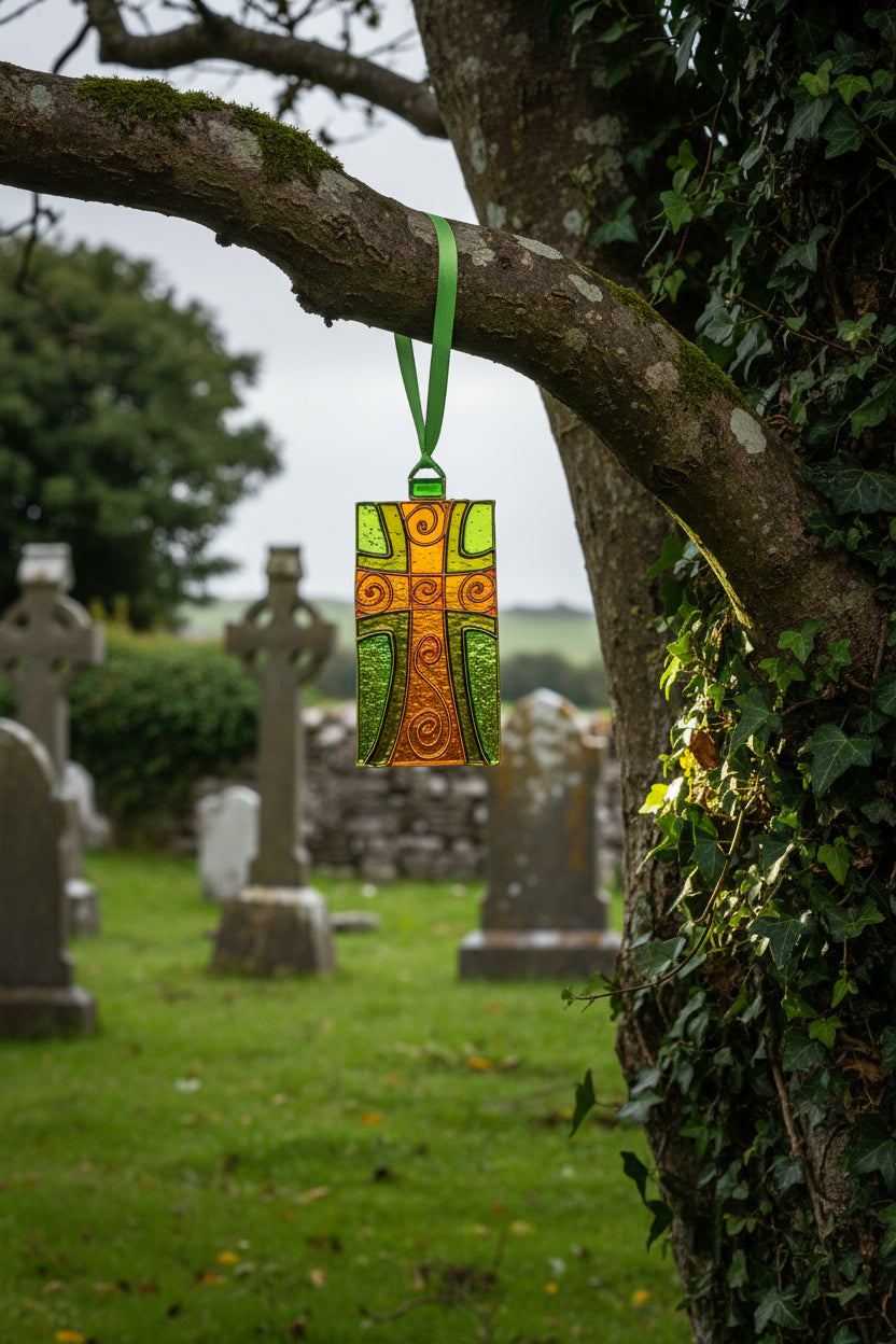 A stained glass hanging sun catcher shaped like a cross, featuring orange and green glass panels with swirling details, attached to a green ribbon. It is displayed in its original packaging with a card labeled 'Visual Instinct' and 'Hanging Sun Catcher' by Irish artist Honor Harte.