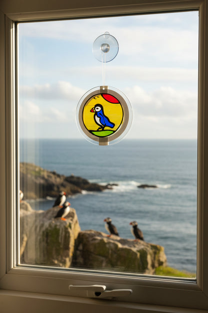 A circular stained glass sun catcher featuring a painted blue bird on a yellow sun over green grass, mounted on a clear stand and shown with its retail card. The card reads 'Sun Catcher "Sticky Beag"' and shows the Visual Instinct brand.