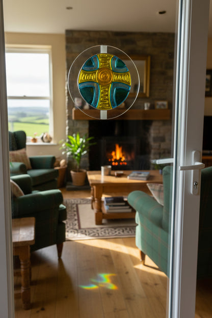 A hand-painted glass sun catcher featuring a Celtic cross design in yellow, blue, and green, shown on its white backing card with the brand Visual Instinct.