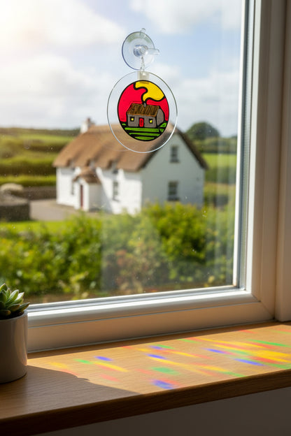 A circular glass ornament labeled 'Sun Catcher Sticky Beag' featuring a hand-painted cottage under a yellow sun on a red and green background, still in its retail packaging with a blue and white header card.