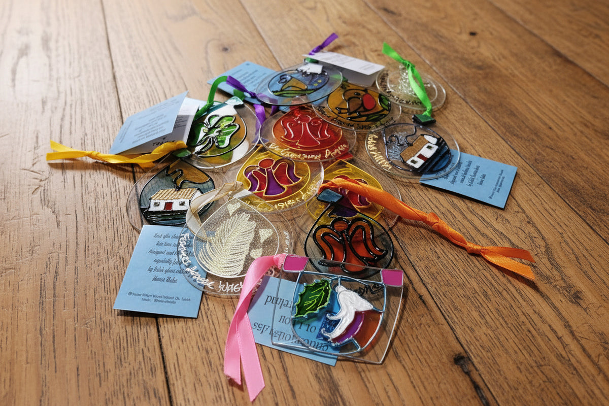 A collection of colorful hand-blown glass ornaments and tags displayed on a light wood surface. The ornaments feature various designs including shamrocks, hearts, a house, and abstract figures in vibrant colors like green, purple, orange, yellow, and blue, with some attached to ribbons and tags.