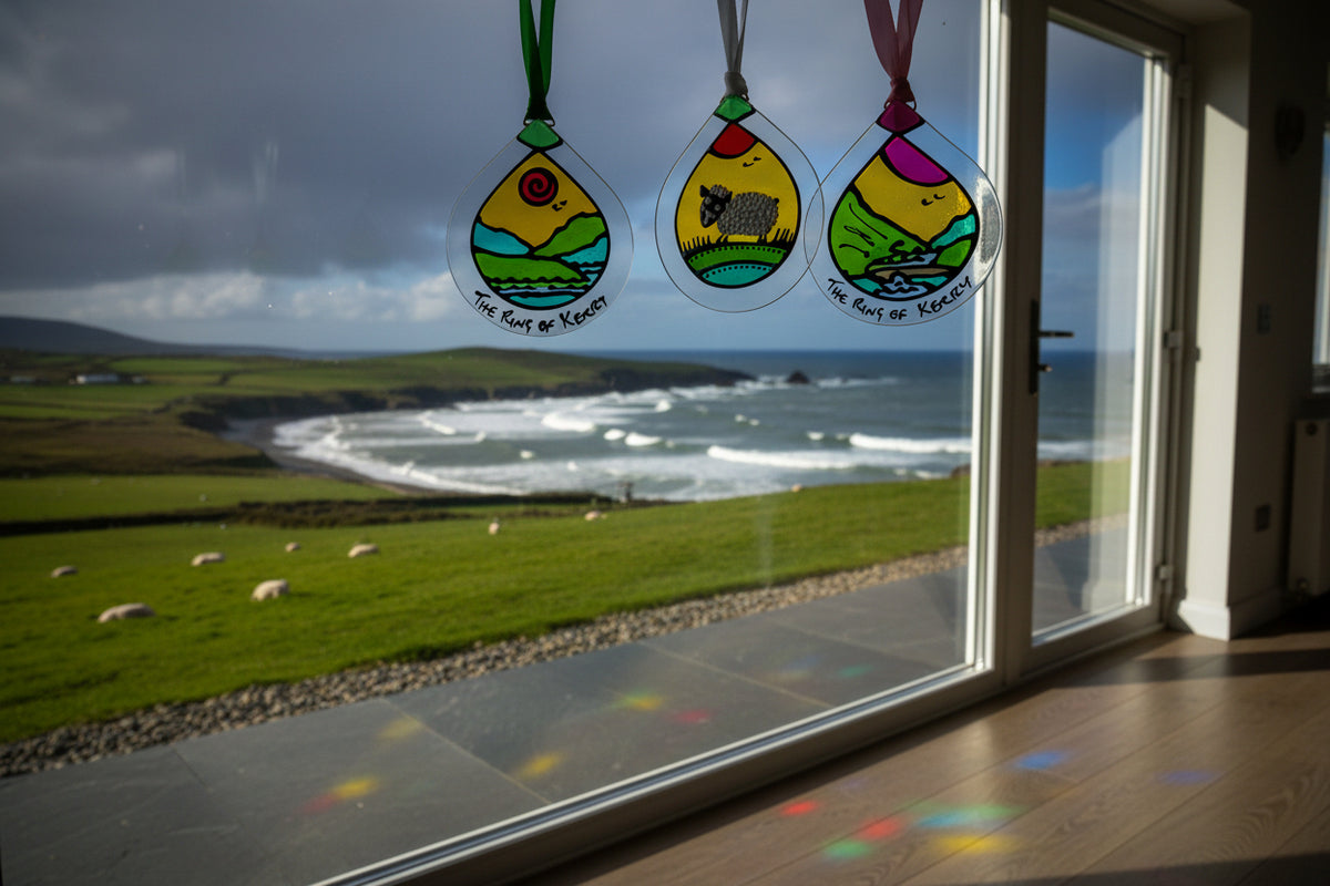 Three hand-painted glass ornaments shaped like teardrops hang from white, green, and pink ribbons. One ornament shows a black-and-white cow in a field, and two others show coastal landscapes with the text 'The Cliffs of Kerney' written along the bottom.