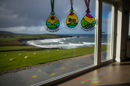 Three hand-painted glass ornaments shaped like teardrops hang from white, green, and pink ribbons. One ornament shows a black-and-white cow in a field, and two others show coastal landscapes with the text 'The Cliffs of Kerney' written along the bottom.