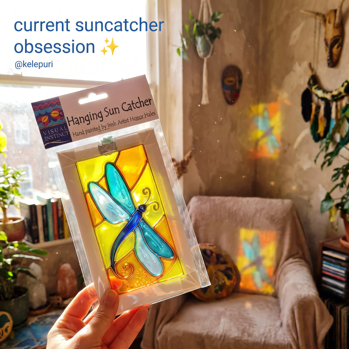 A hand holds a packaged stained glass hanging sun catcher shaped like a dragonfly, featuring blue and teal wings with a yellow and orange background. The package label reads 'Hanging Sun Catcher' and shows the Visual Aspects brand.