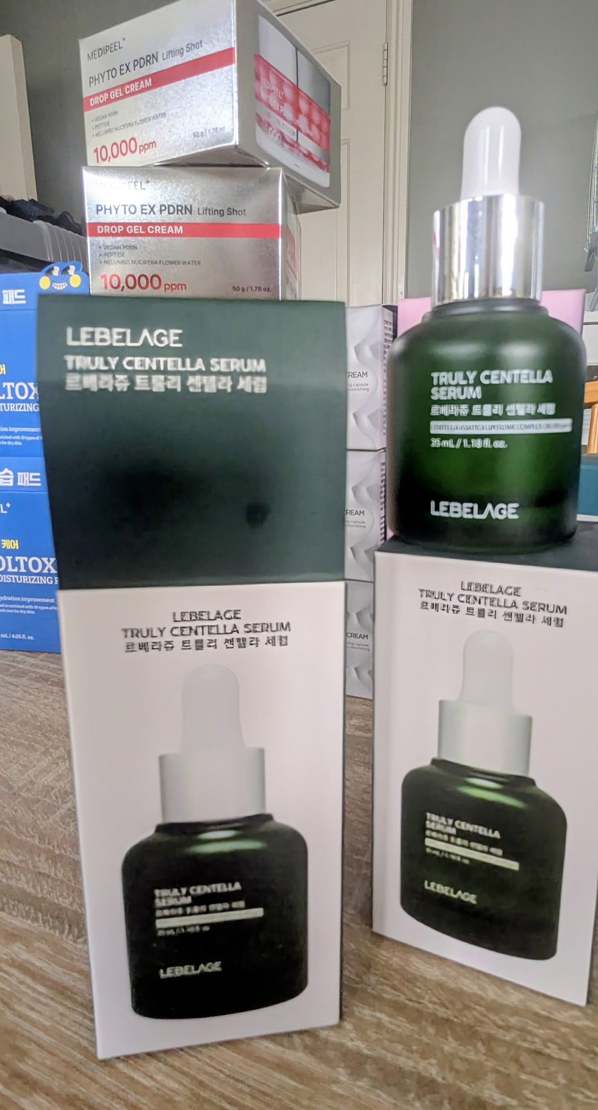 LEBELAGE Truly Centella Serum shown with its green and white retail box, a dark green bottle with a silver dropper cap, and two additional product boxes stacked in the background on a tabletop.