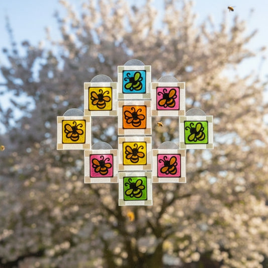 Eleven small square bee suncatchers in blue, pink, orange, yellow and green, arranged on a window against blossom