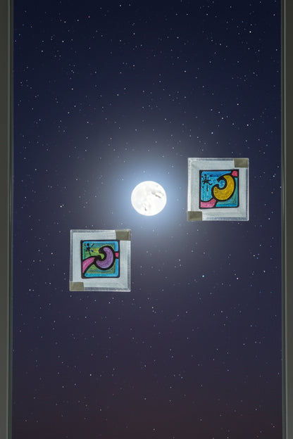 Two small square crescent moon and star suncatchers on a window beneath a full moon and starry sky